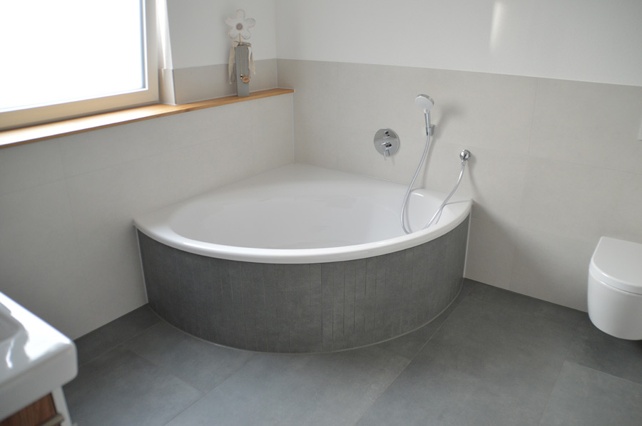 Einbau-Eckbadewanne mit grauen Fliesen verkleidet und Unterputzarmaturen