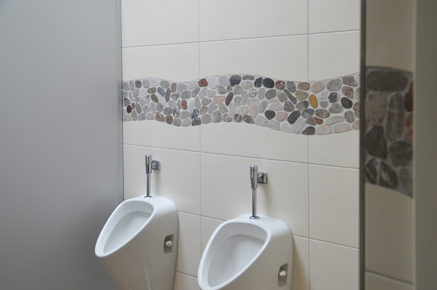 Zwei Urinale an weißer Wandfliese mit dekorativer Flussstein-Bordüre
