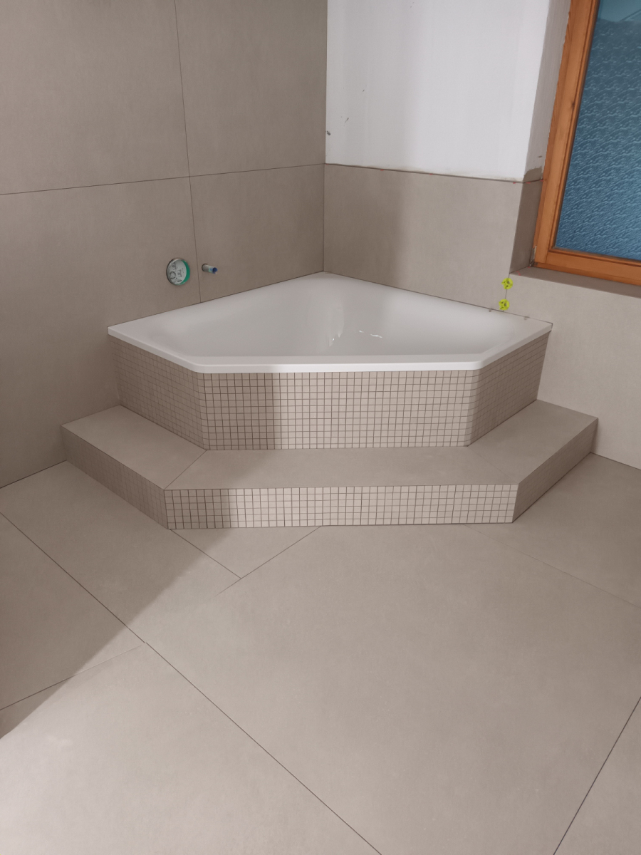 Weiße Eckbadewanne auf zweistufigem Podest mit beiger Mosaikfliesen-Verkleidung und großformatigen Bodenfliesen